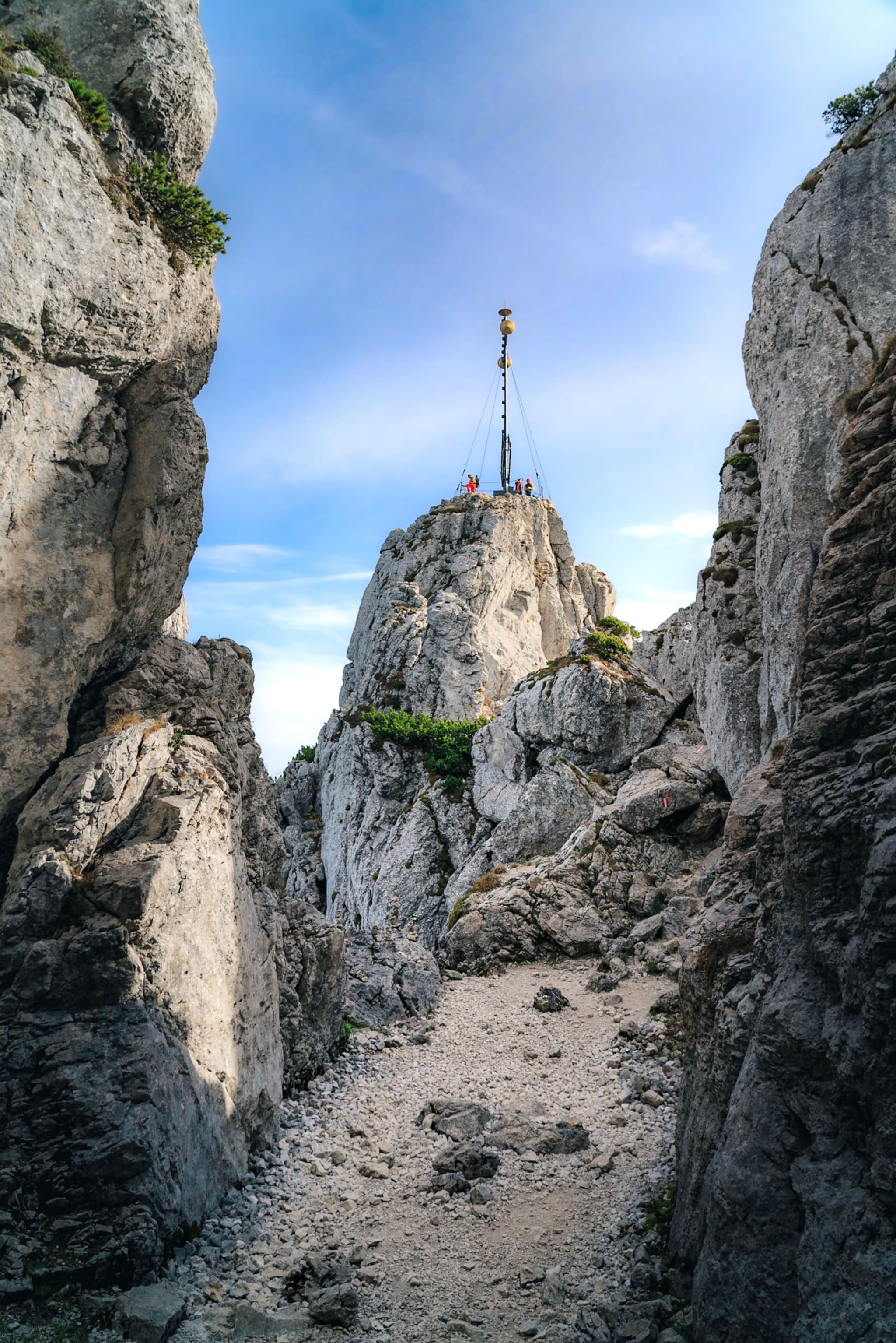 A narrow rocky path leads to a peak with a cross at the top, surrounded by large rock formations under a clear blue sky