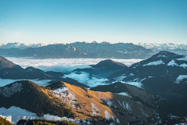 High mountain range with snow-dusted peaks and sunlit ridges rising above a sea of clouds under a clear blue sky