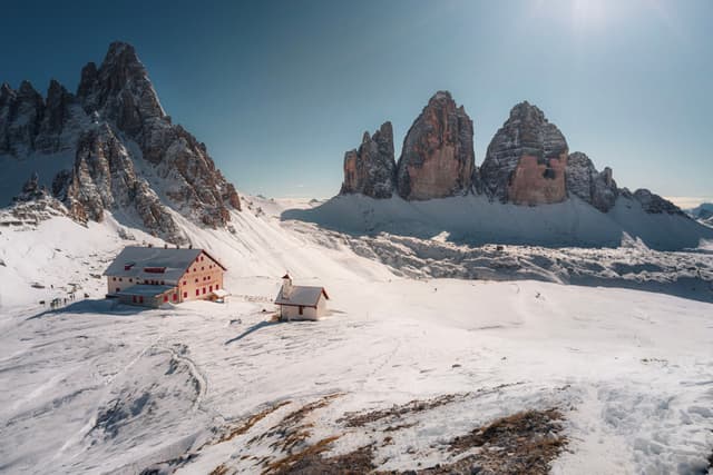 Snow-covered alpine lodges beneath towering rocky peaks under a clear blue sky