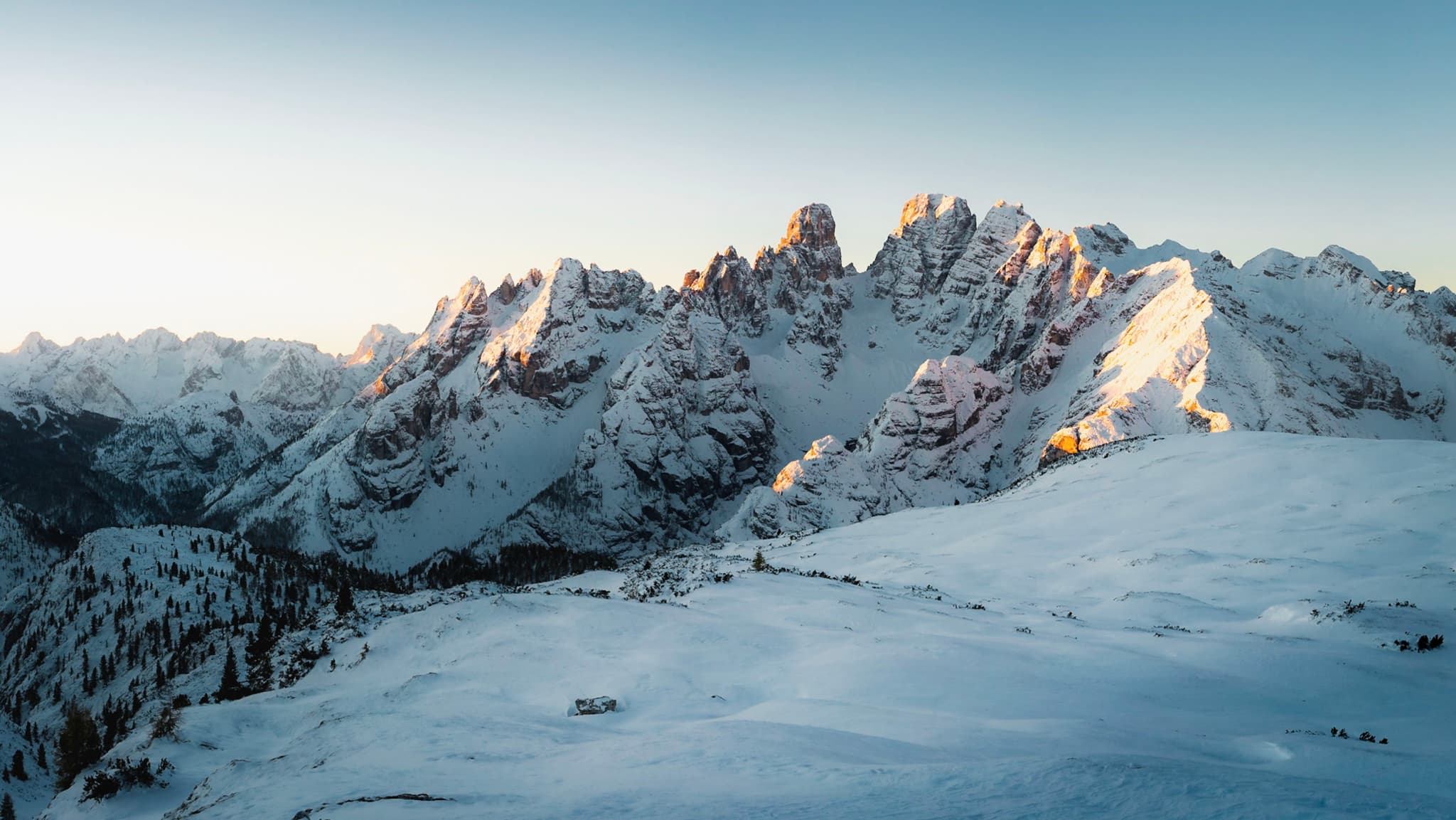 Snow-covered mountain range at sunrise with a small illuminated campsite on a ridge