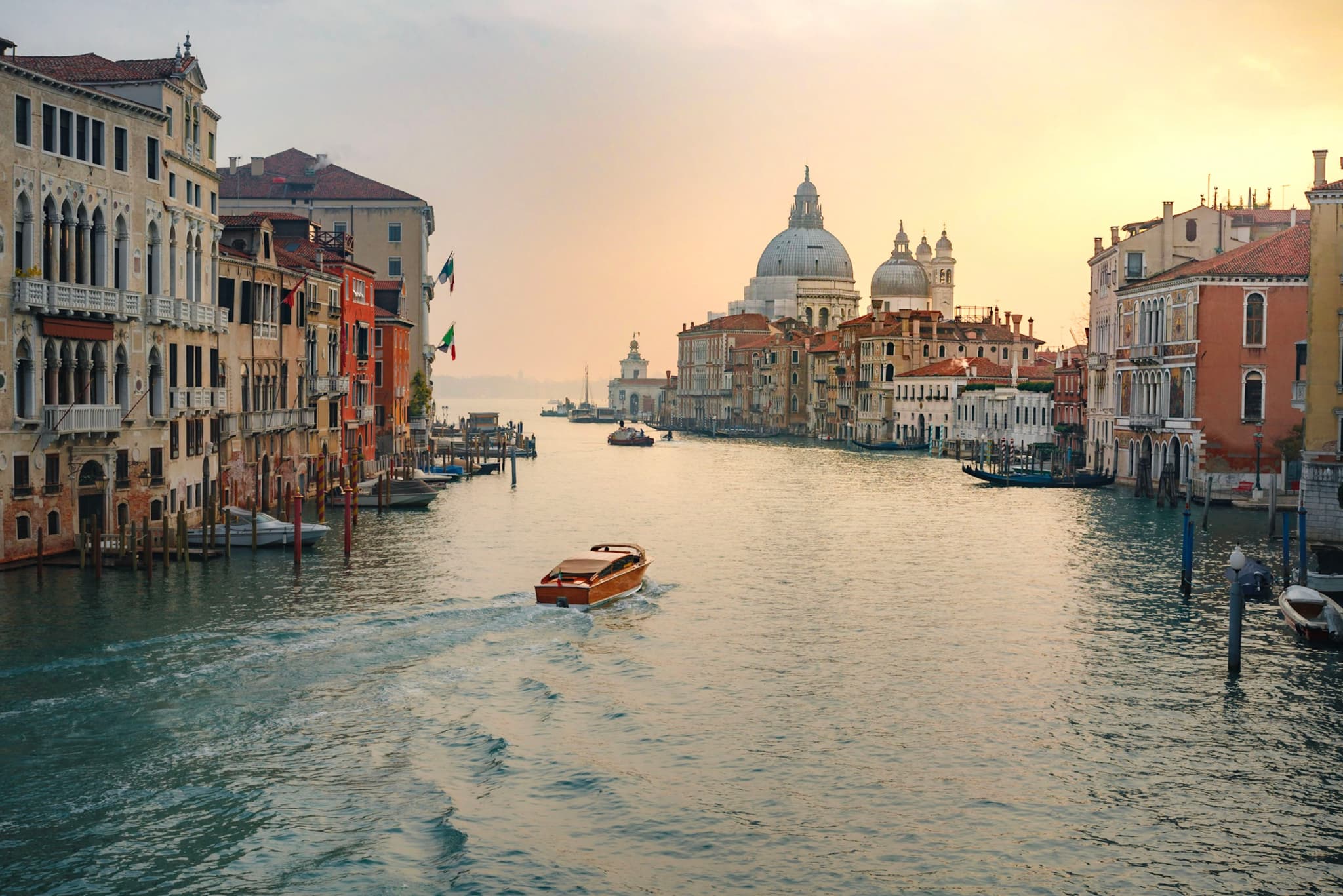 A scenic canal in Venice with boats navigating the waterway, surrounded by historic buildings and a prominent domed structure in the background under a warm, glowing sky