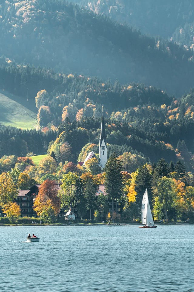 Serene lake with small sailboat and rowboat in the foreground, backed by a forested hillside and a white church steeple rising above autumn trees