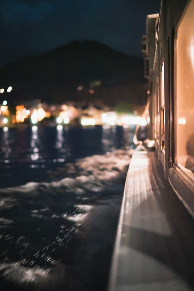 Boat gliding through dark harbor waters at night with distant shoreline lights and a silhouetted hill in the background