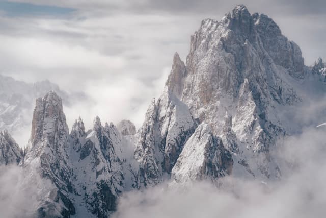 Snow-covered jagged mountain peaks rise through a layer of clouds, creating a dramatic and serene landscape