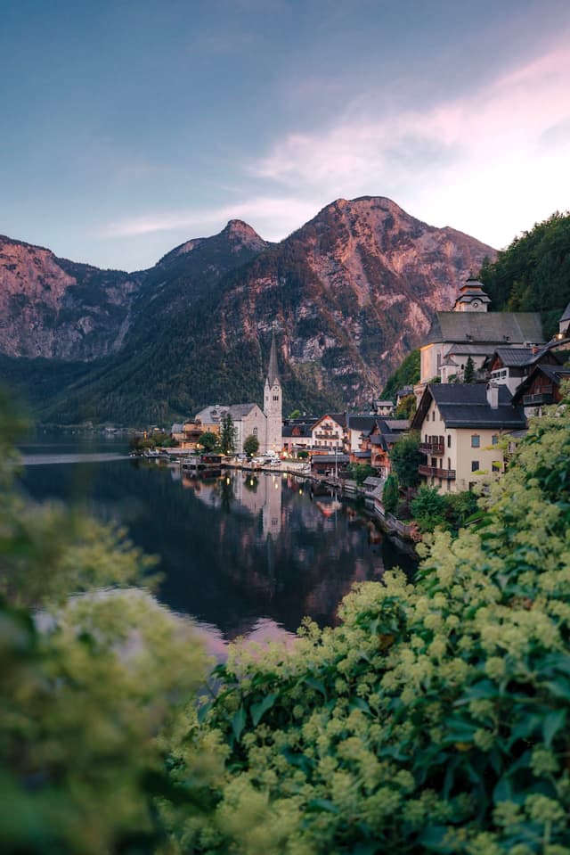 A picturesque lakeside village with charming buildings, a prominent church spire, and majestic mountains in the background, surrounded by lush greenery