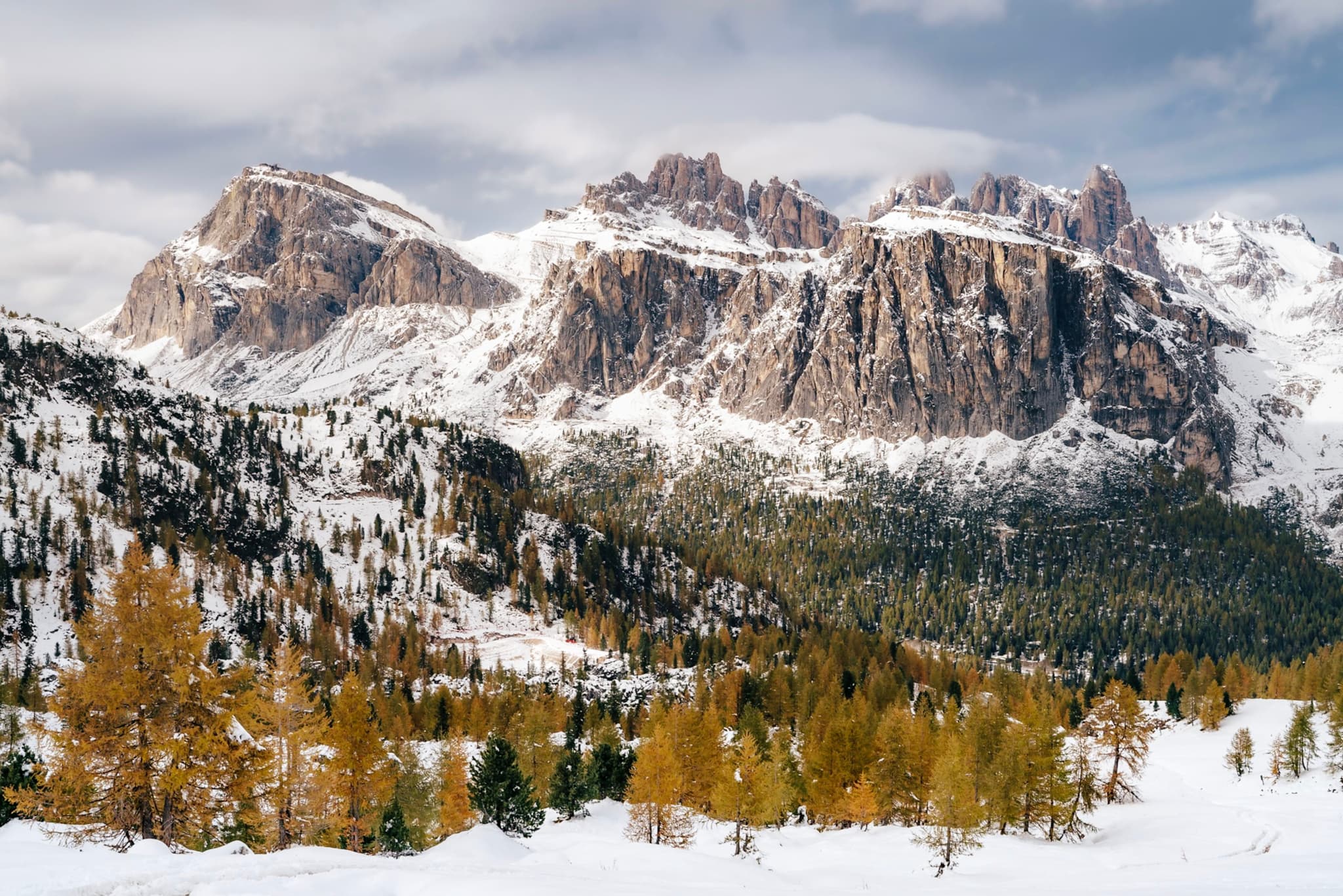 A snowy mountain range with rugged peaks, surrounded by a forest of evergreen and golden-leaved trees under a partly cloudy sky