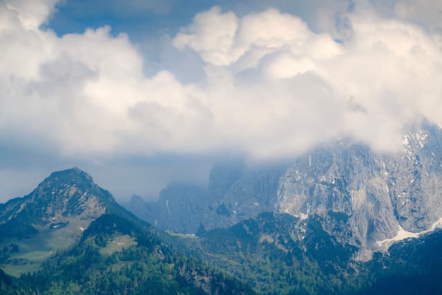 A mountainous landscape with peaks partially obscured by clouds, featuring lush greenery in the foreground