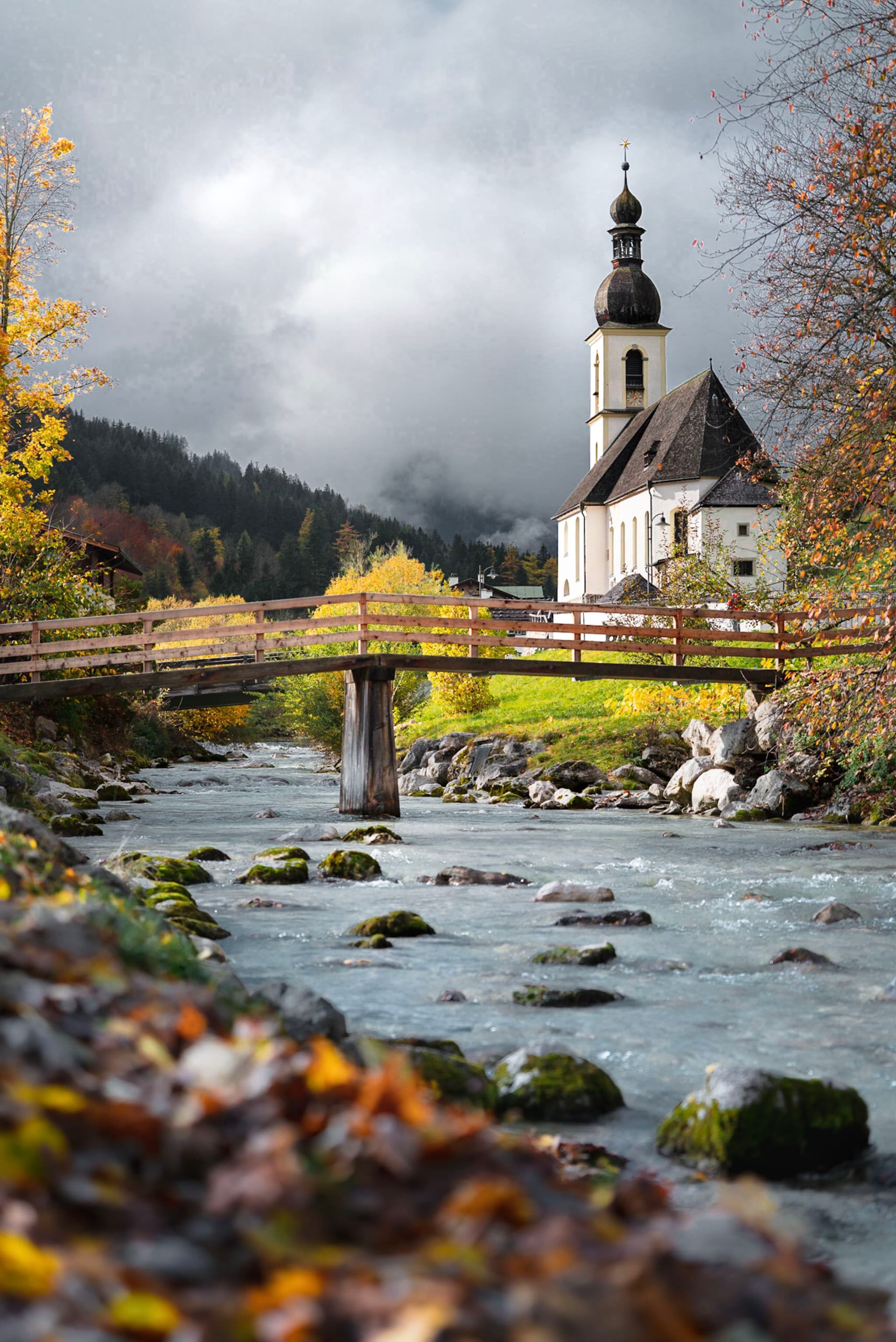 A picturesque scene featuring a church with a tall steeple, set against a backdrop of cloudy skies and mountains. A wooden bridge crosses a flowing river, surrounded by autumn foliage