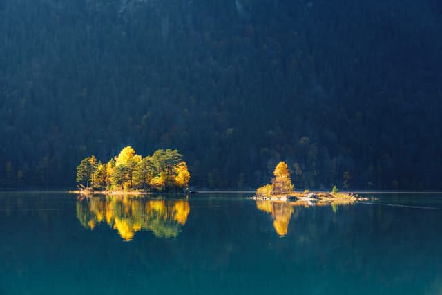 A serene lake with small islands covered in autumn-colored trees, reflecting on the calm water, set against a backdrop of dark forested hills