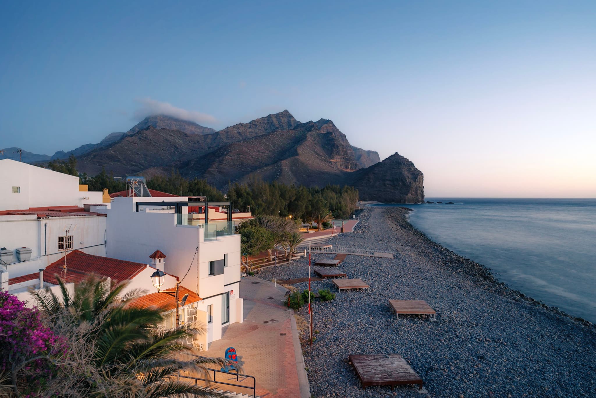 Quiet seaside village with white houses and palm trees beside a pebbled beach at sunrise, backed by steep mountains along a calm sea