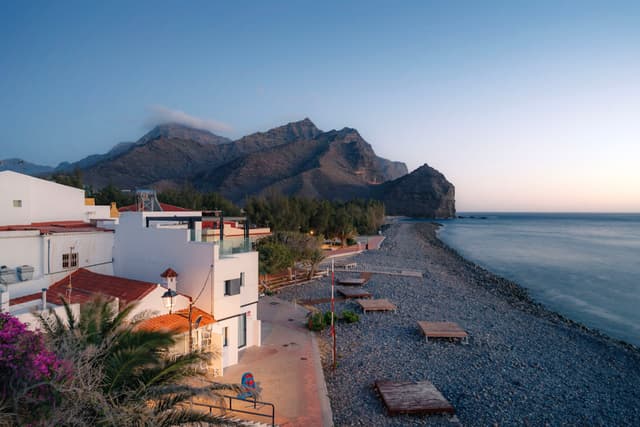 Quiet seaside village with white houses and palm trees beside a pebbled beach at sunrise, backed by steep mountains along a calm sea