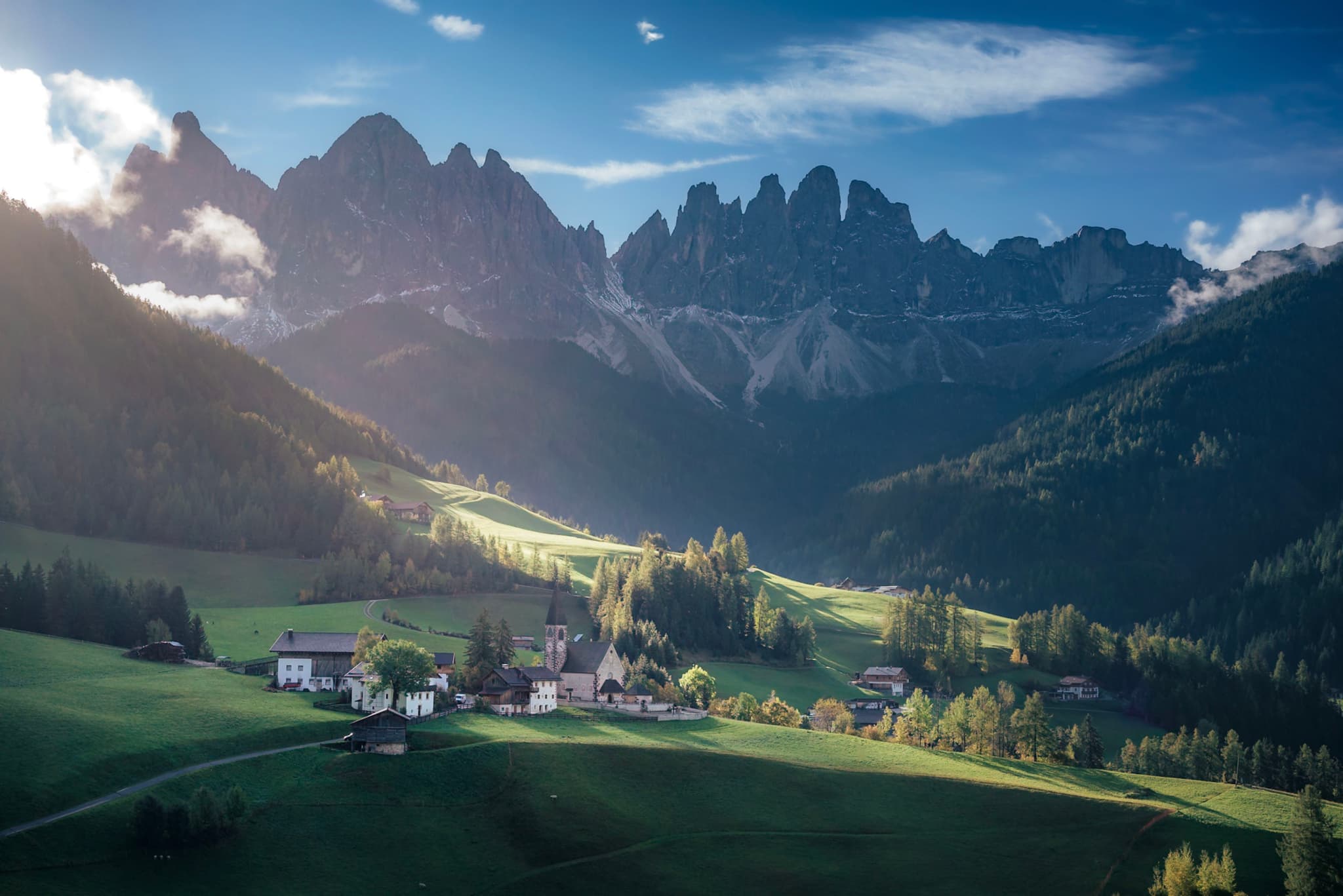 A picturesque village nestled in a lush green valley, surrounded by dramatic mountain peaks under a partly cloudy sky