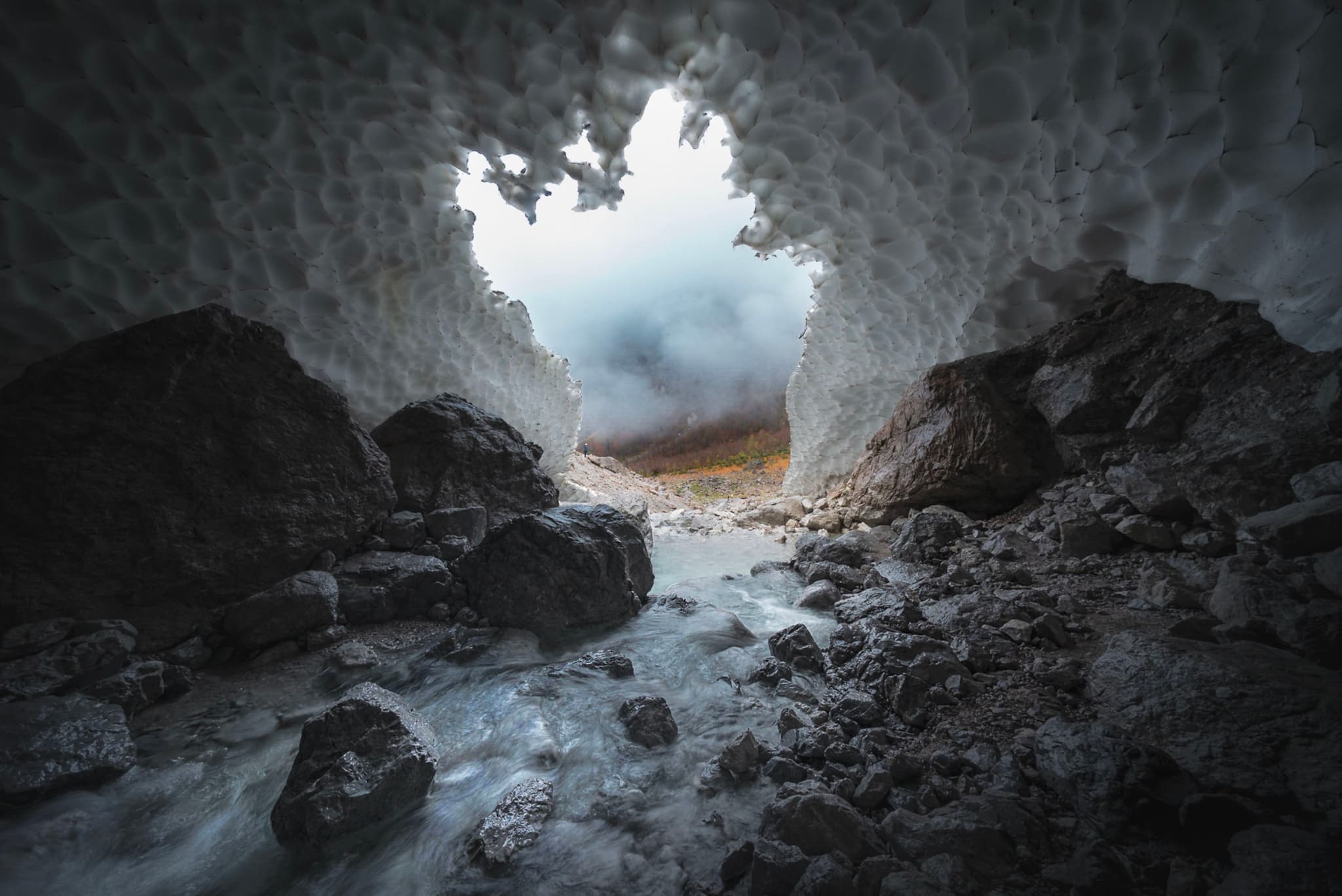 Misty light pours through an opening in a snow-covered cave, illuminating rocky ground and a trickling stream leading out into a foggy landscape