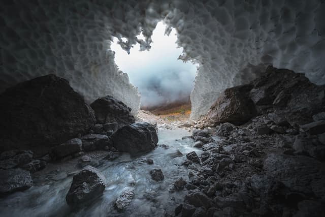 Misty light pours through an opening in a snow-covered cave, illuminating rocky ground and a trickling stream leading out into a foggy landscape