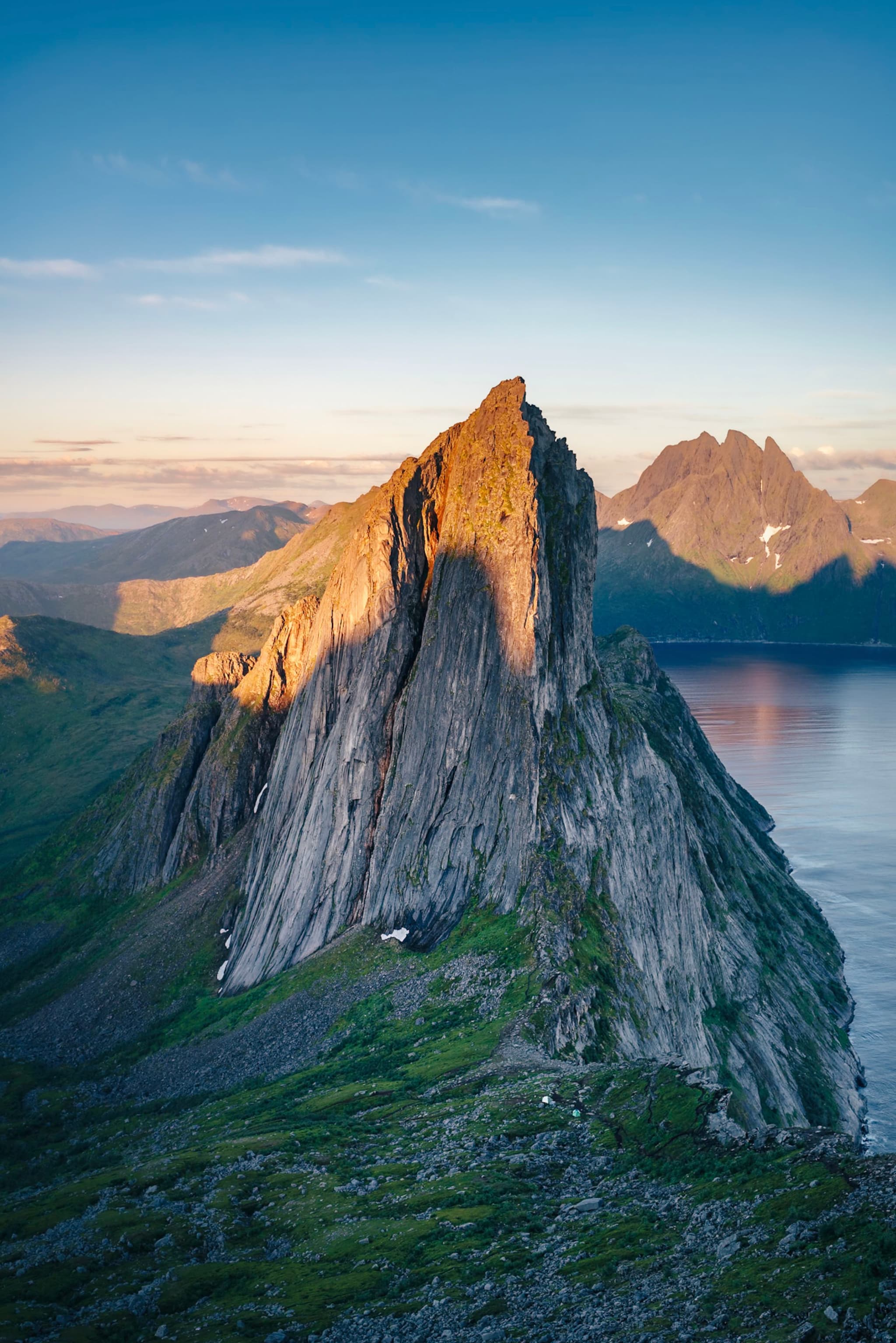 A dramatic, jagged mountain peak bathed in warm sunlight, surrounded by lush green terrain and overlooking a calm body of water with distant mountains in the background