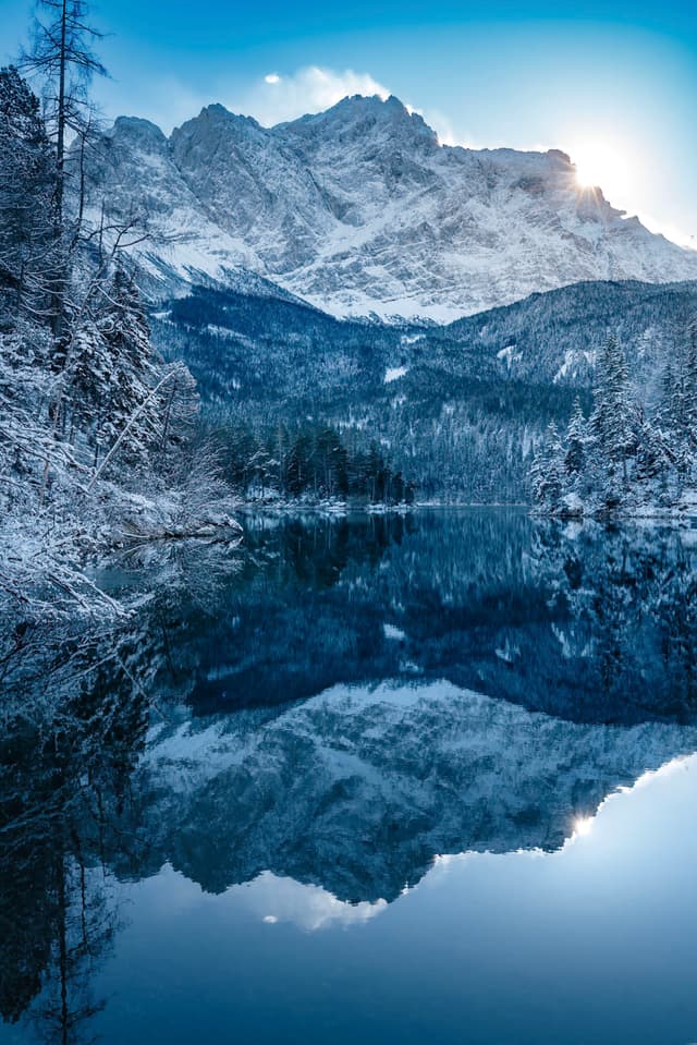 A serene winter landscape with snow-covered mountains reflected in a calm lake, surrounded by frosty trees under a clear blue sky