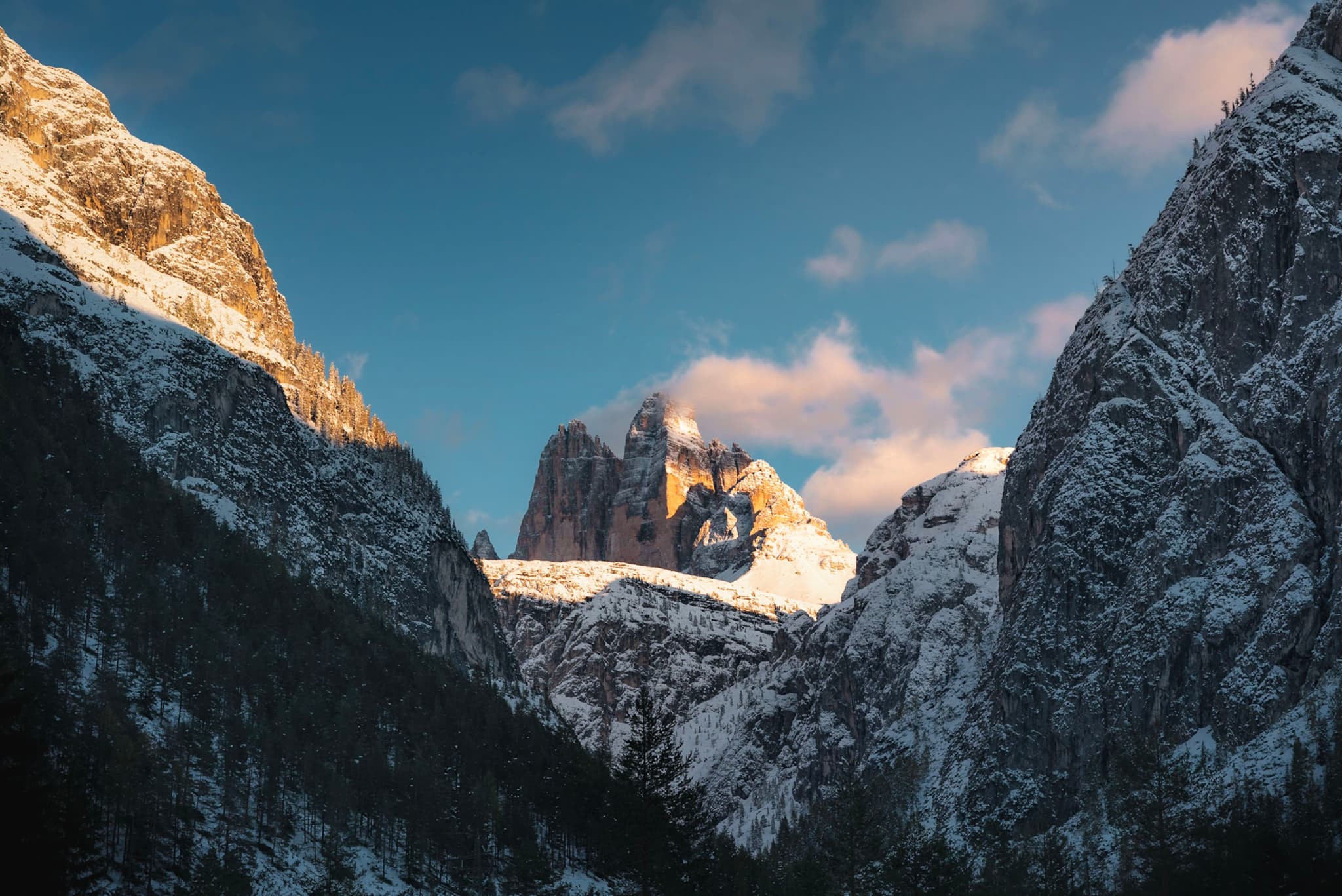 Snow-covered mountains framing a sunlit central peak under a soft pink-cloud sunset sky