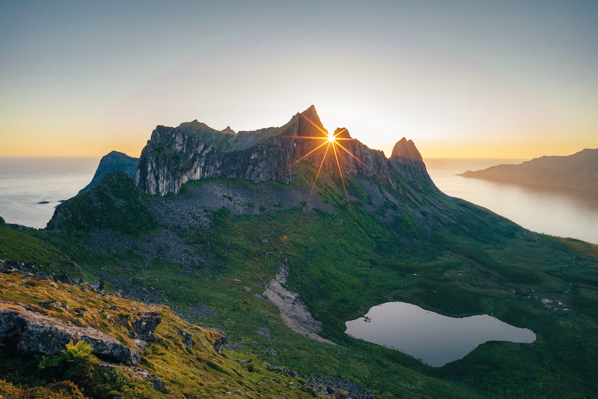 A stunning mountain landscape at sunrise, with the sun peeking over jagged peaks, casting a warm glow over lush green terrain and a small lake in the foreground