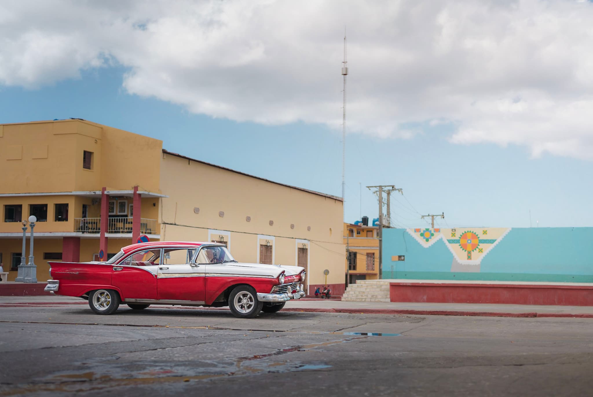 Red and white classic car parked on a wide empty street beside pastel-colored Cuban buildings under a cloudy sky