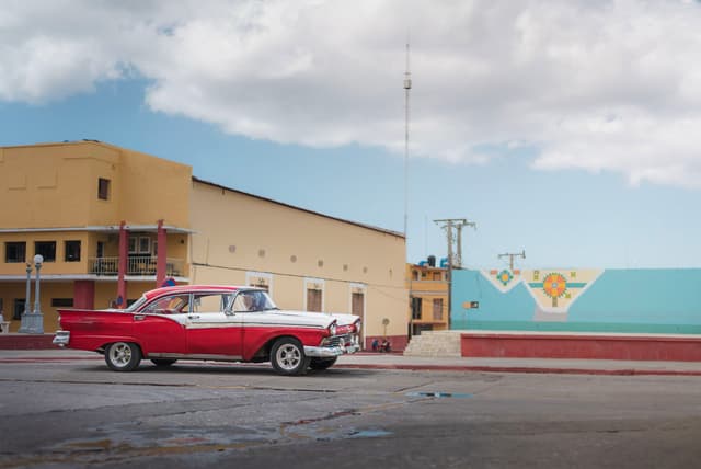 Red and white classic car parked on a wide empty street beside pastel-colored Cuban buildings under a cloudy sky