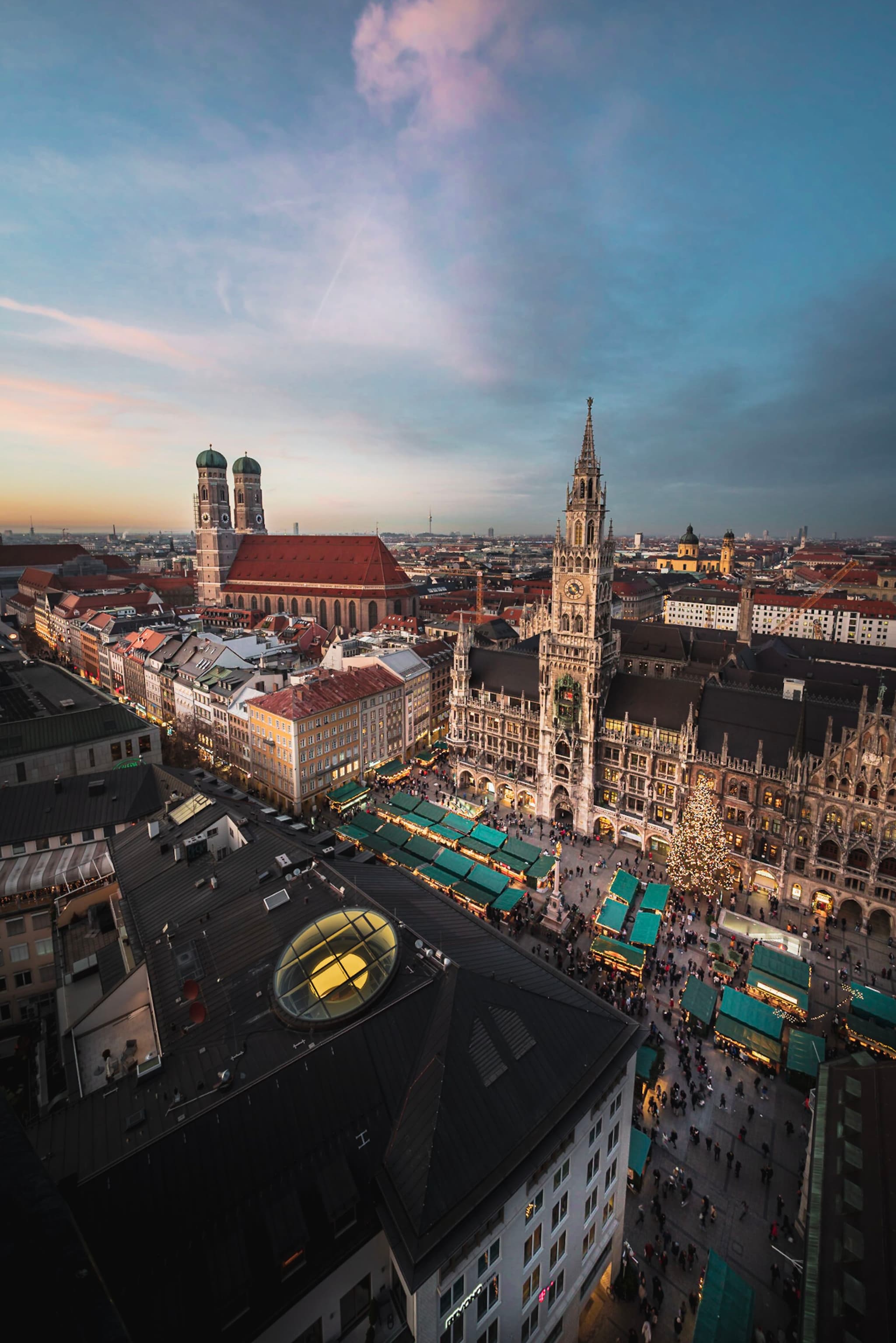 Historic European city square at dusk with a tall gothic-style clock tower, busy market stalls, and warm lights illuminating crowded streets