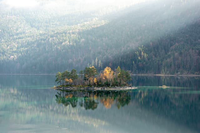 A small, tree-covered island is reflected in a calm lake, surrounded by misty mountains