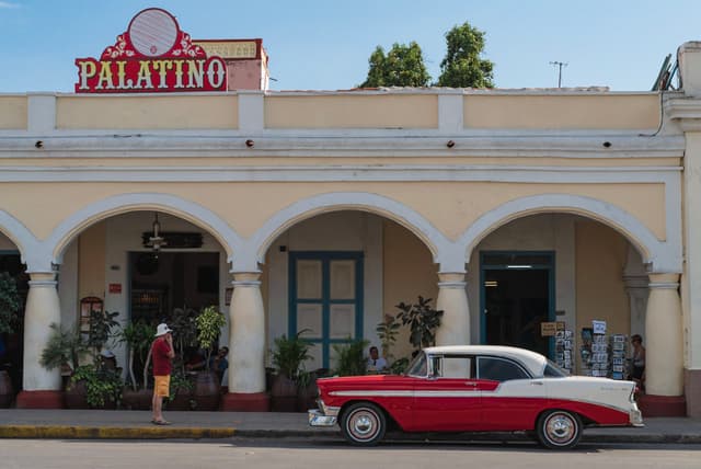 Vintage red-and-white American car parked in front of the Palatino arcade facade with arches, plants, and a pedestrian on the sidewalk