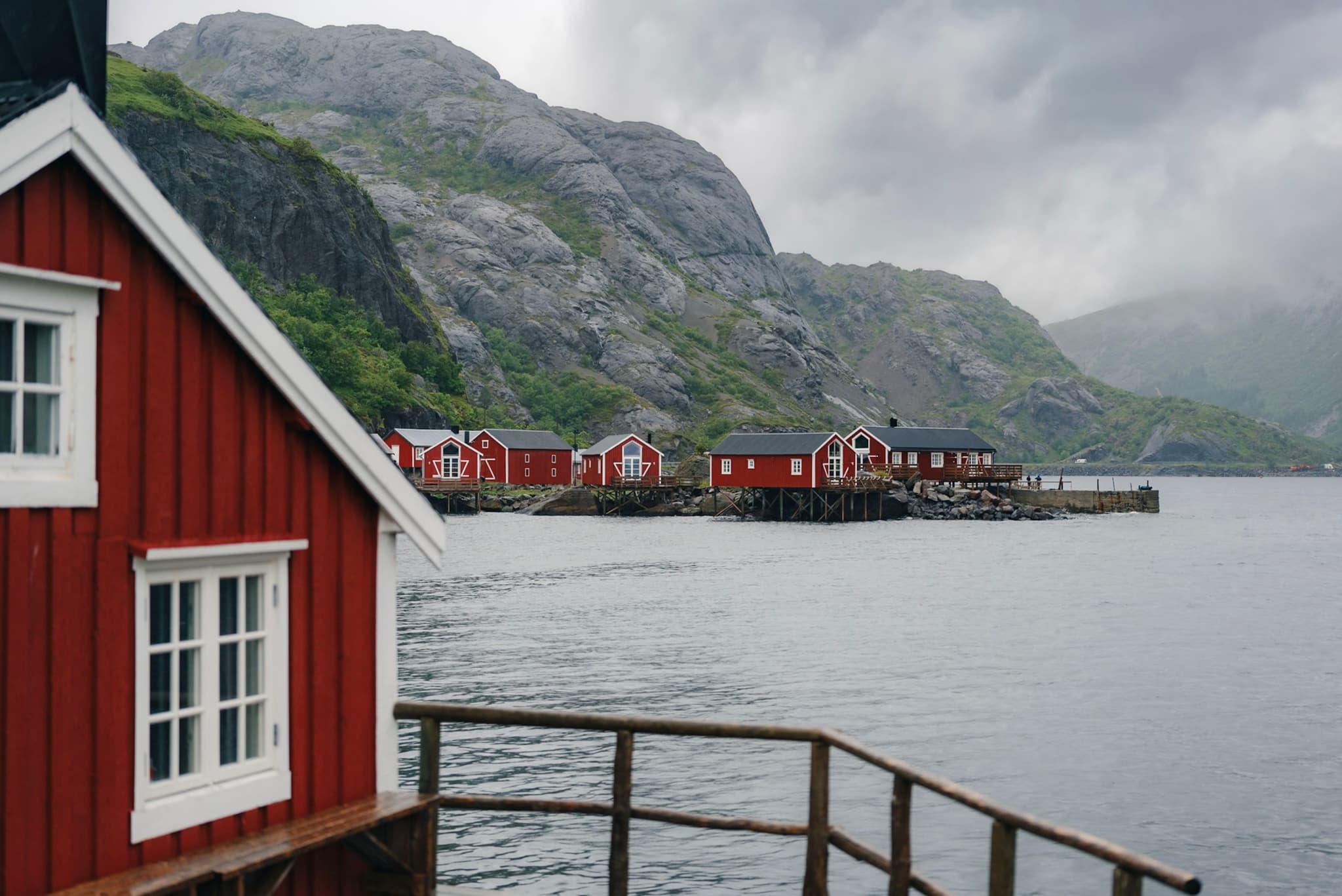 Red wooden houses by a calm waterfront, surrounded by rocky hills under a cloudy sky