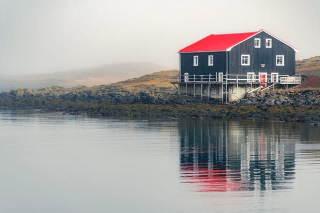 A house with a red roof and dark walls sits by a calm body of water, with its reflection visible The background is misty and serene