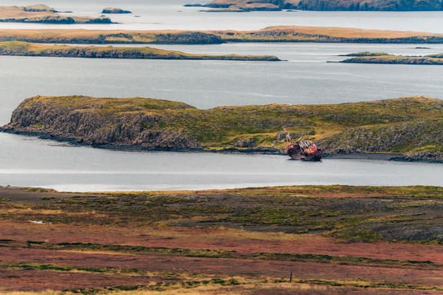 Rusting shipwreck resting in a narrow channel between low, grassy islands and layered coastal shallows