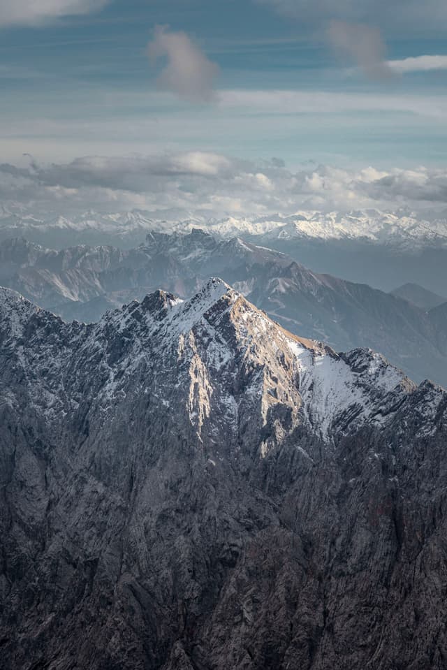 Sunlit snow-dusted mountain peak rising sharply above layered ranges under a cloudy sky