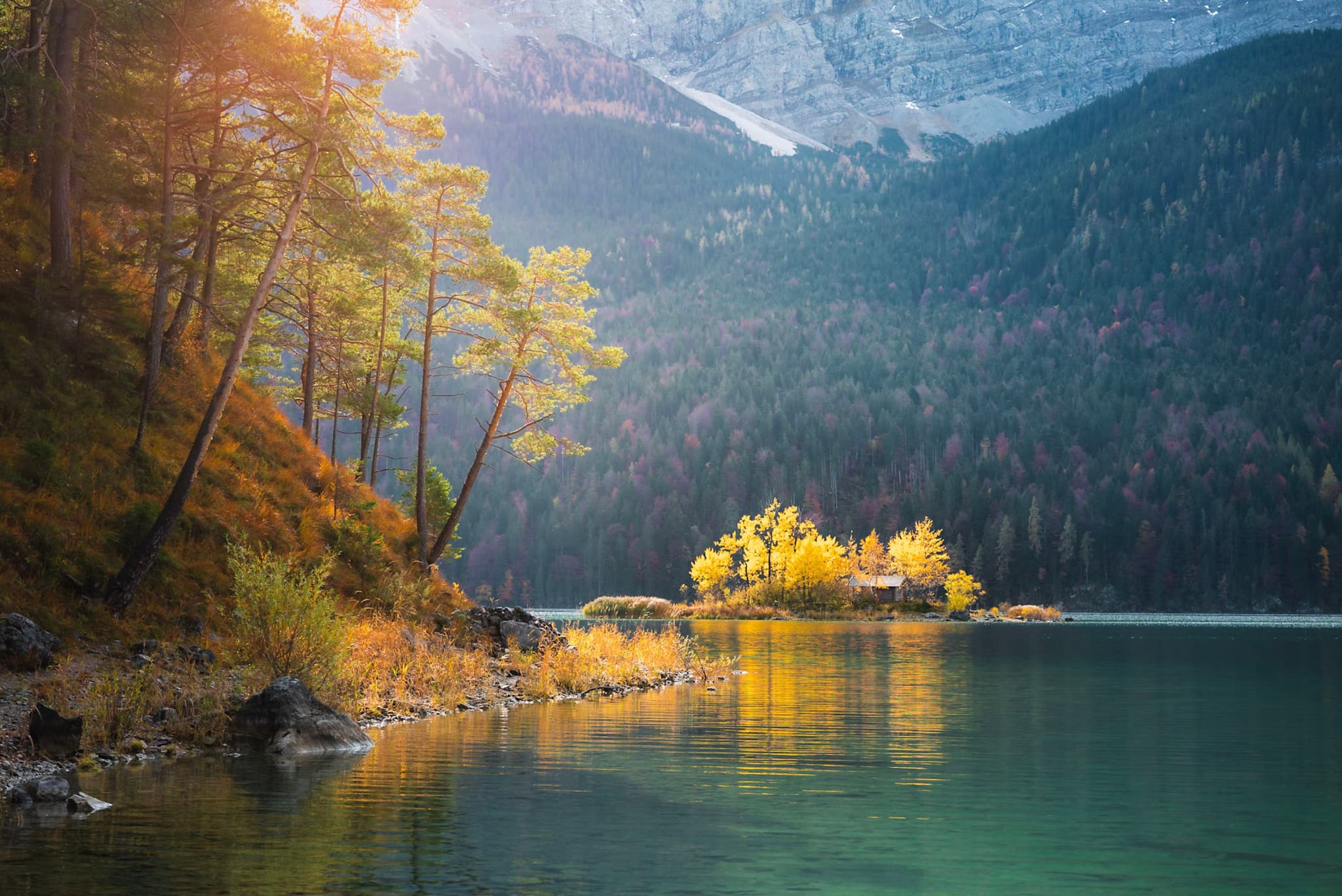 Tranquil mountain lake with a small sunlit forested island reflecting on calm turquoise water, surrounded by autumn trees and distant snowy peaks