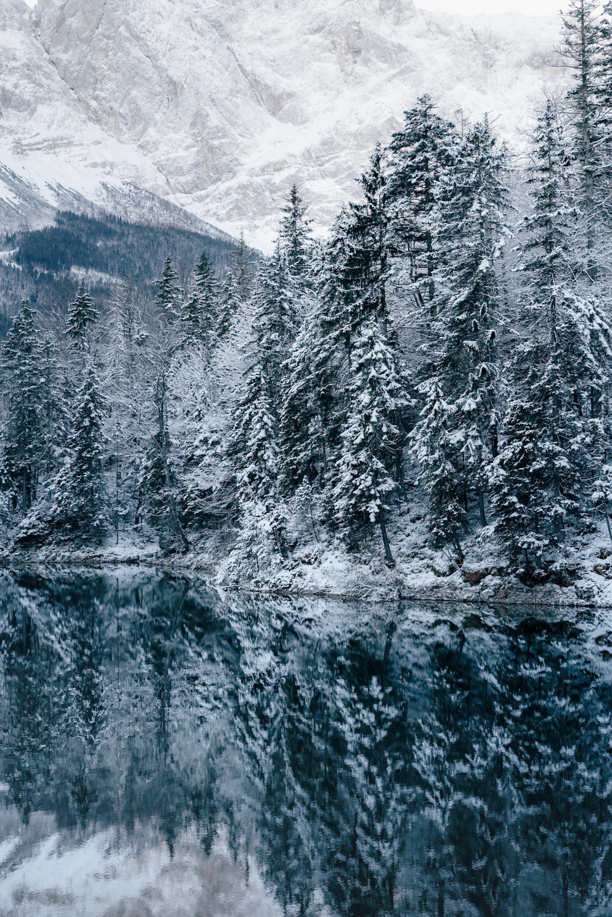 A serene winter landscape with snow-covered trees reflected in a calm, icy lake, set against a backdrop of snowy mountains