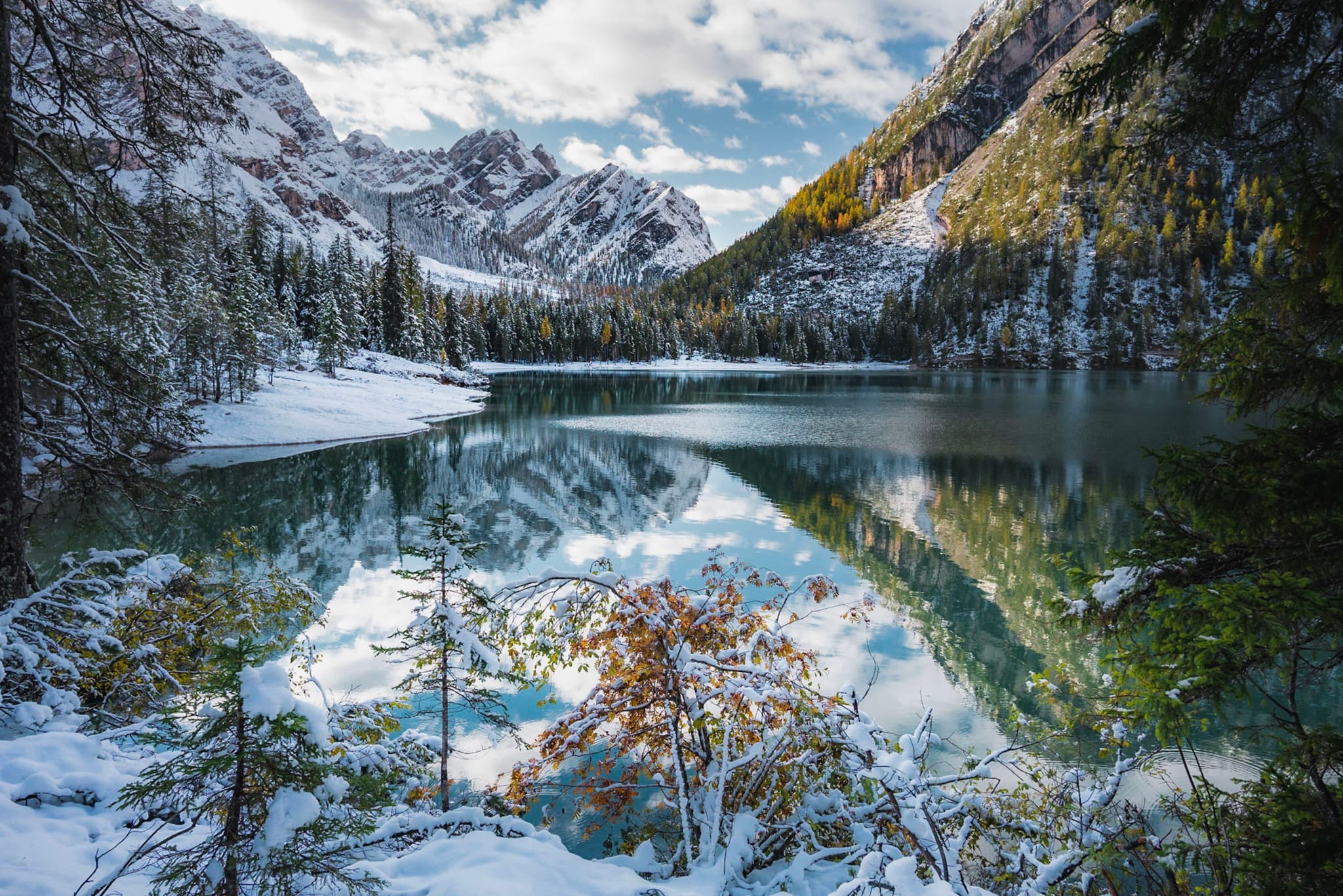 Snow-dusted alpine lake reflecting surrounding evergreen forests and rugged mountain peaks under a partly cloudy sky