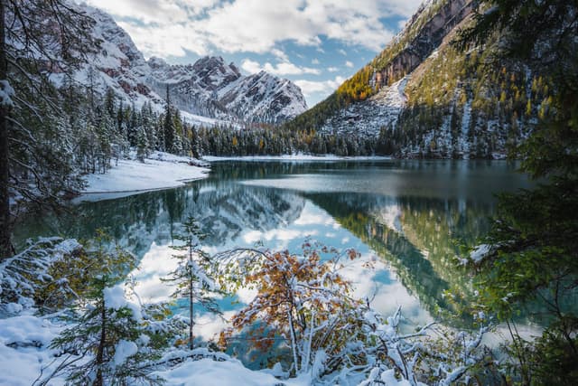 Snow-dusted alpine lake reflecting surrounding evergreen forests and rugged mountain peaks under a partly cloudy sky