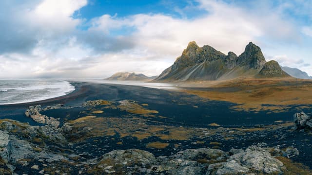 Dramatic jagged mountains rising above a black sand beach and rocky shoreline under a partly cloudy sky with waves rolling in from the ocean
