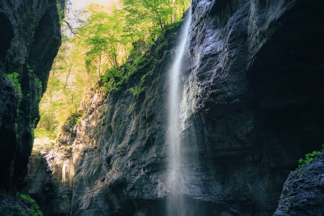 Tall waterfall dropping between steep rock walls into a narrow canyon with sunlit trees above