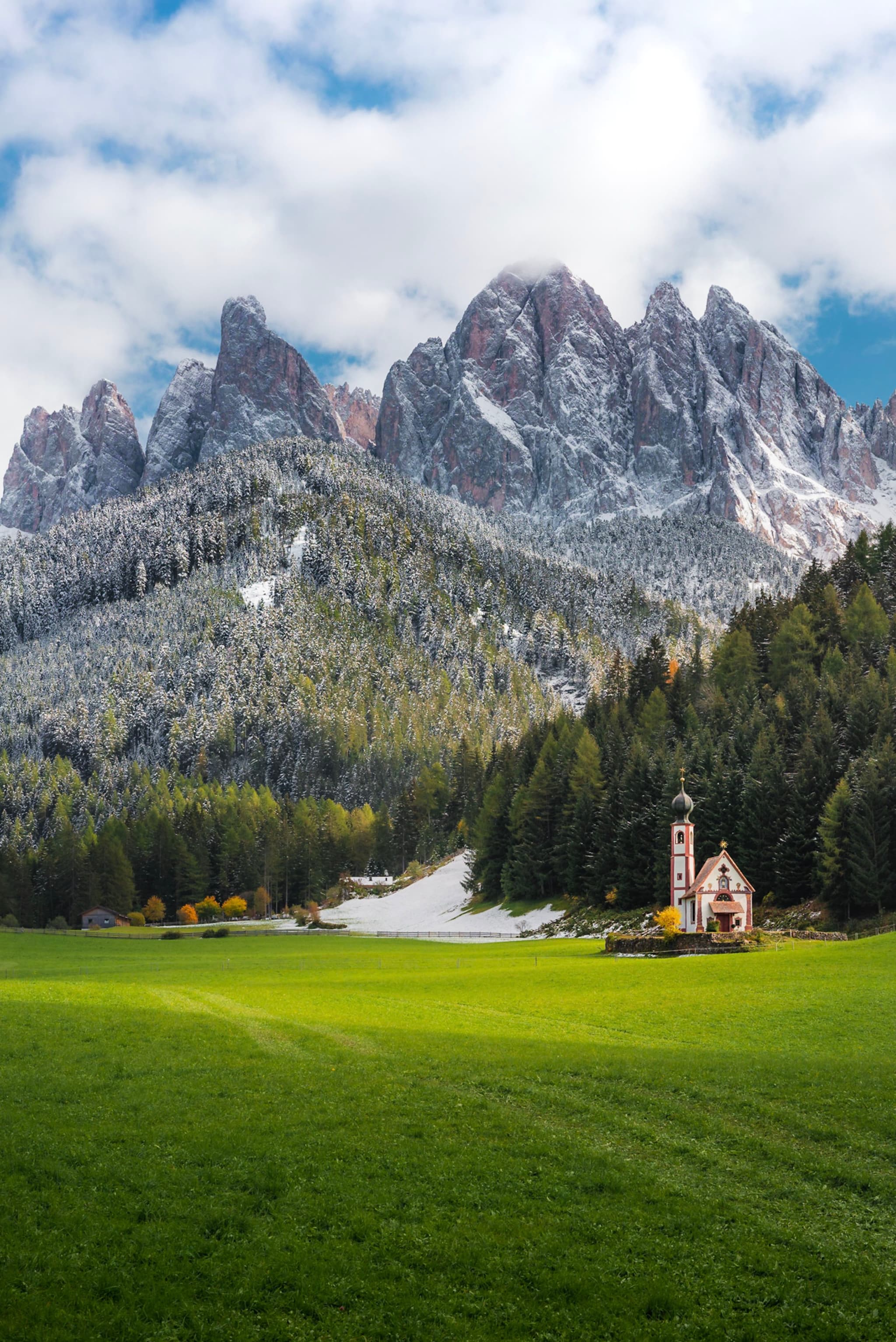 Tiny country church beside dark evergreen forest at the base of towering snow-dusted mountains and a bright green meadow under a partly cloudy sky