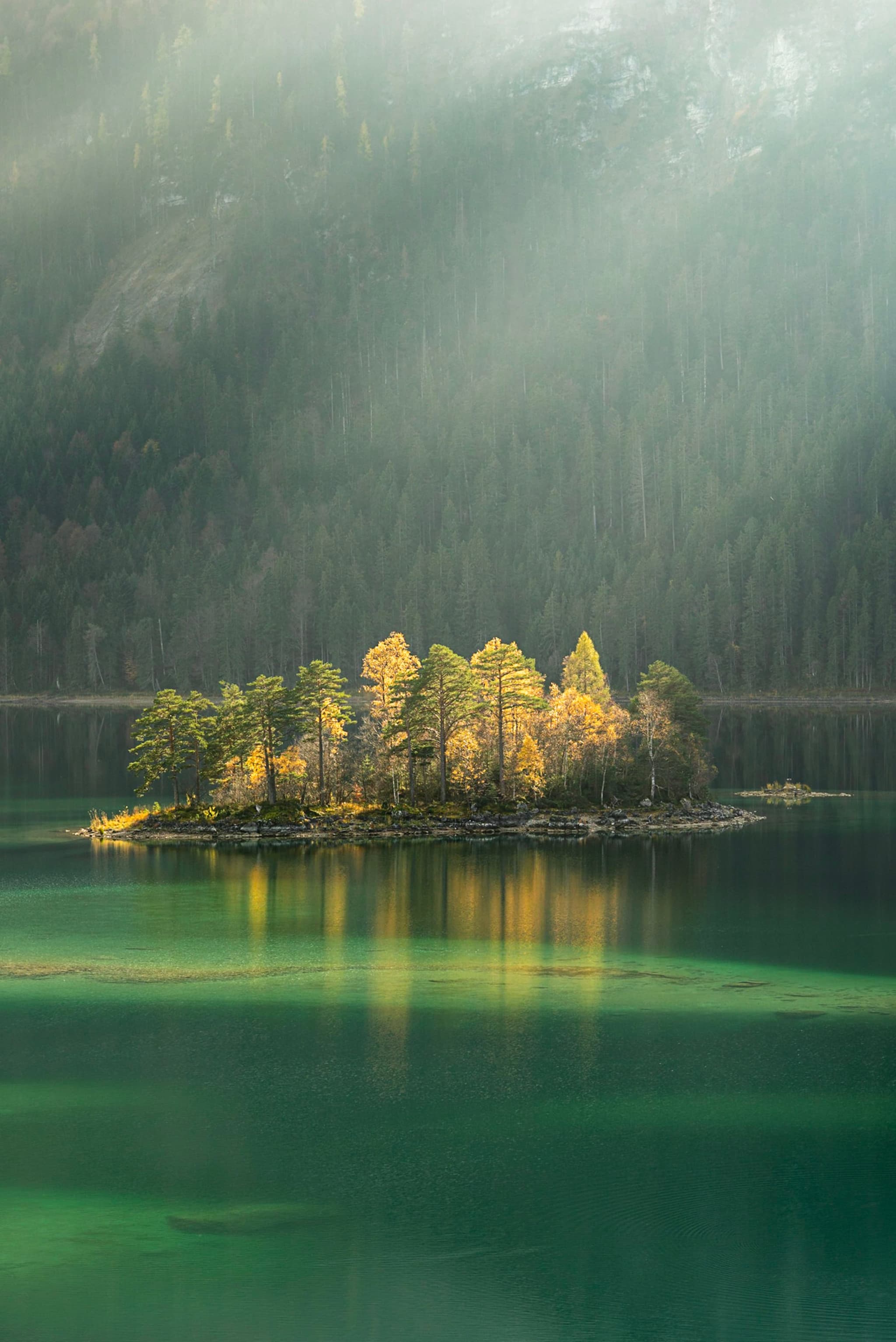 A small, tree-covered island surrounded by calm, green water with sunlight streaming through a misty atmosphere