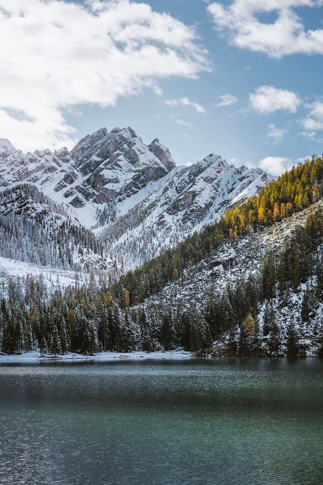 Snow-dusted pine slopes and rugged alpine peaks reflected in a calm mountain lake under a partly cloudy sky
