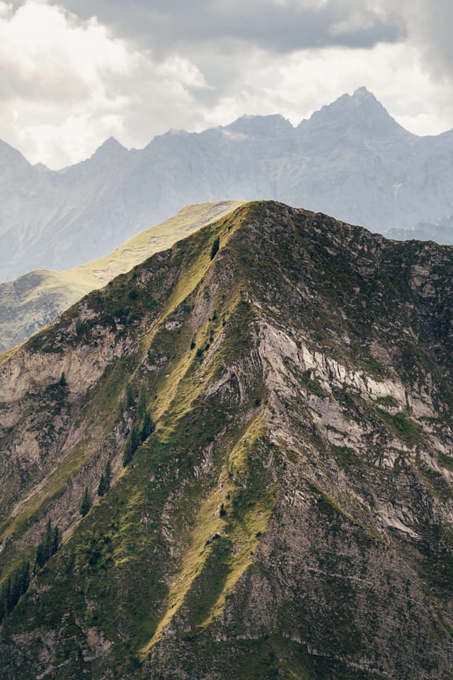 Steep green mountain ridge rising toward distant jagged peaks under a cloudy sky