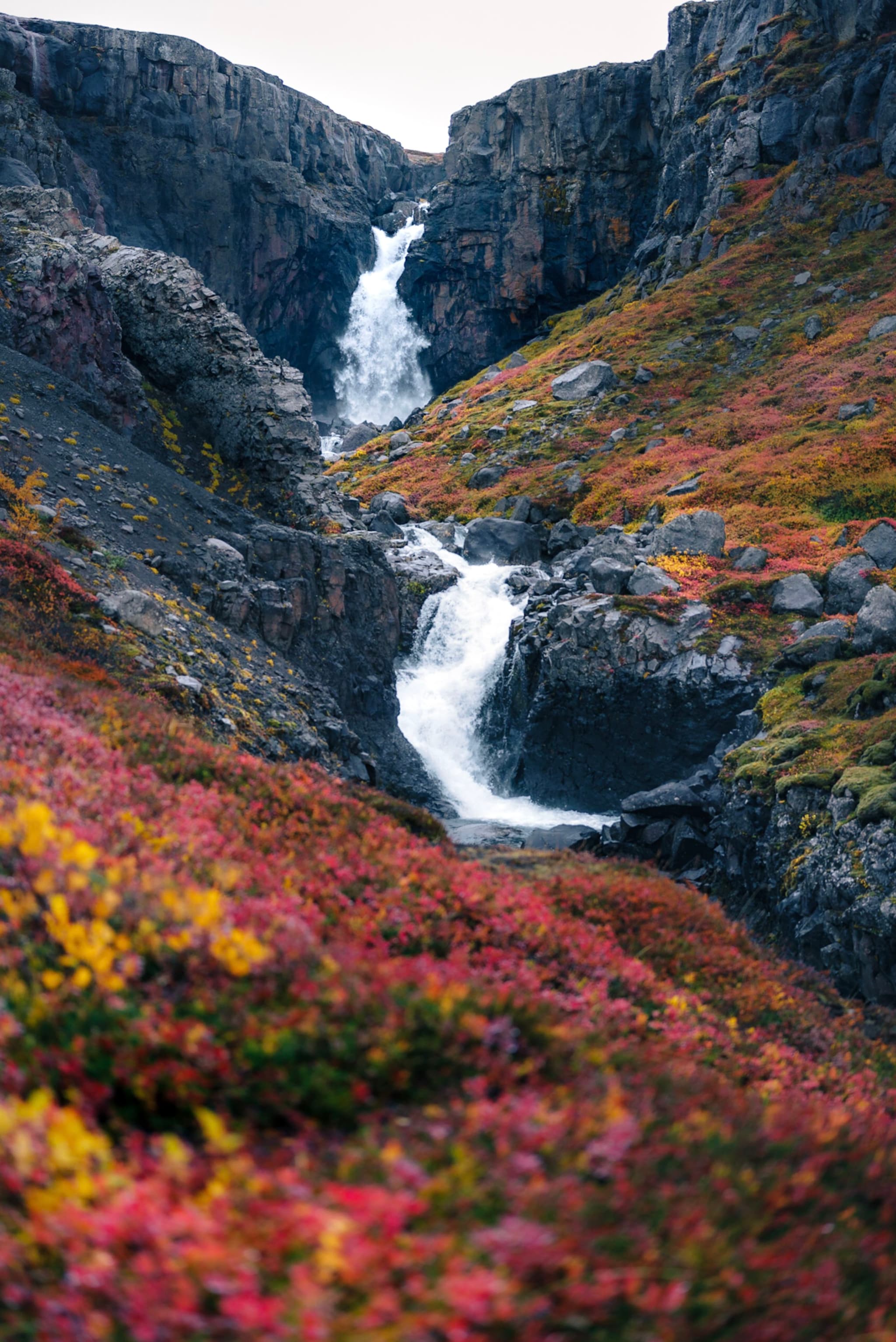 Multi-tiered waterfall cascading through a rocky canyon framed by vibrant autumn foliage in red, yellow, and orange tones