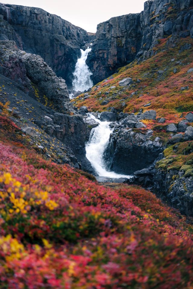 Multi-tiered waterfall cascading through a rocky canyon framed by vibrant autumn foliage in red, yellow, and orange tones