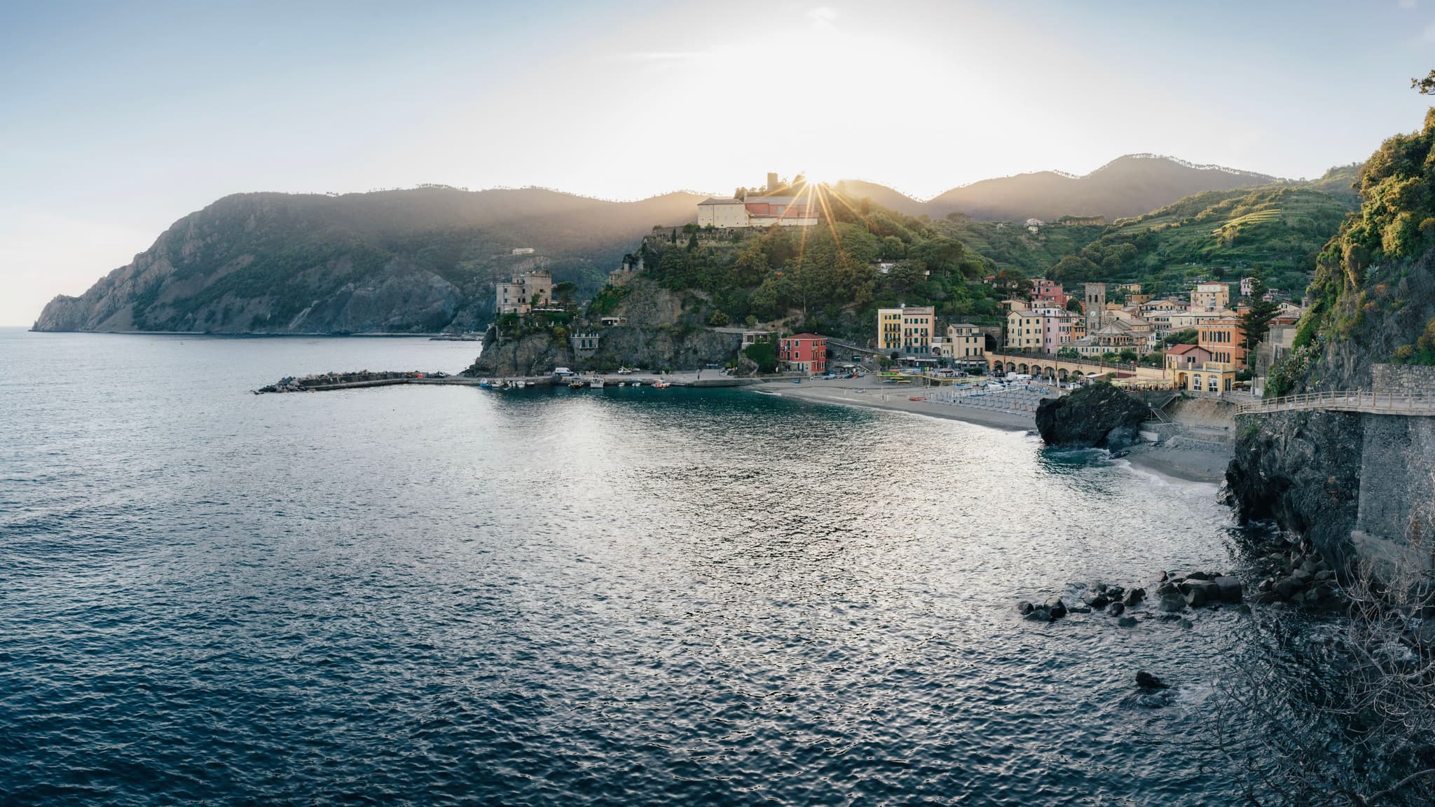 A coastal town with colorful buildings nestled between rugged cliffs and a calm sea, illuminated by the setting sun