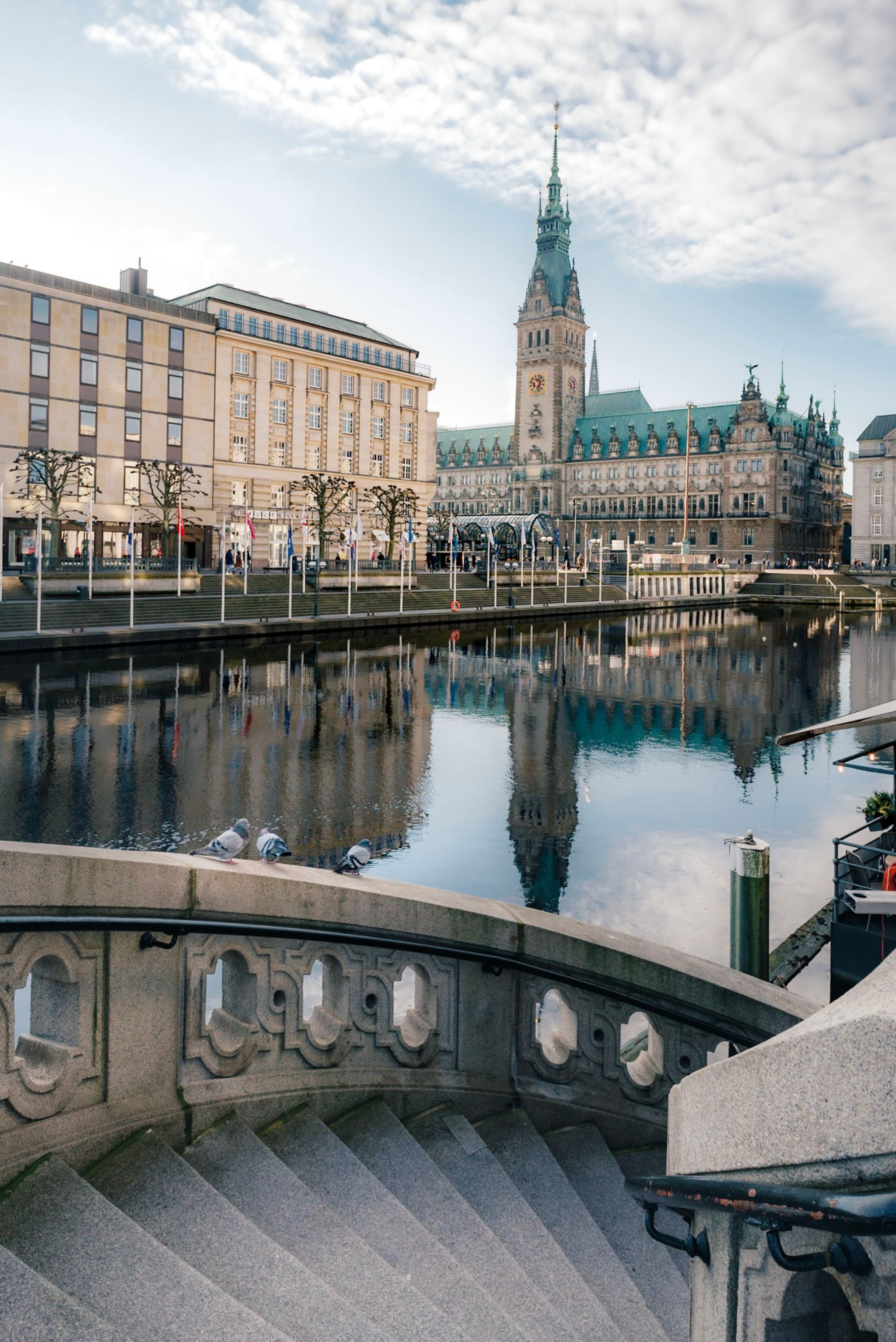 Stone staircase leading to a calm canal reflecting historic waterfront buildings and a tall clock tower under a partly cloudy sky