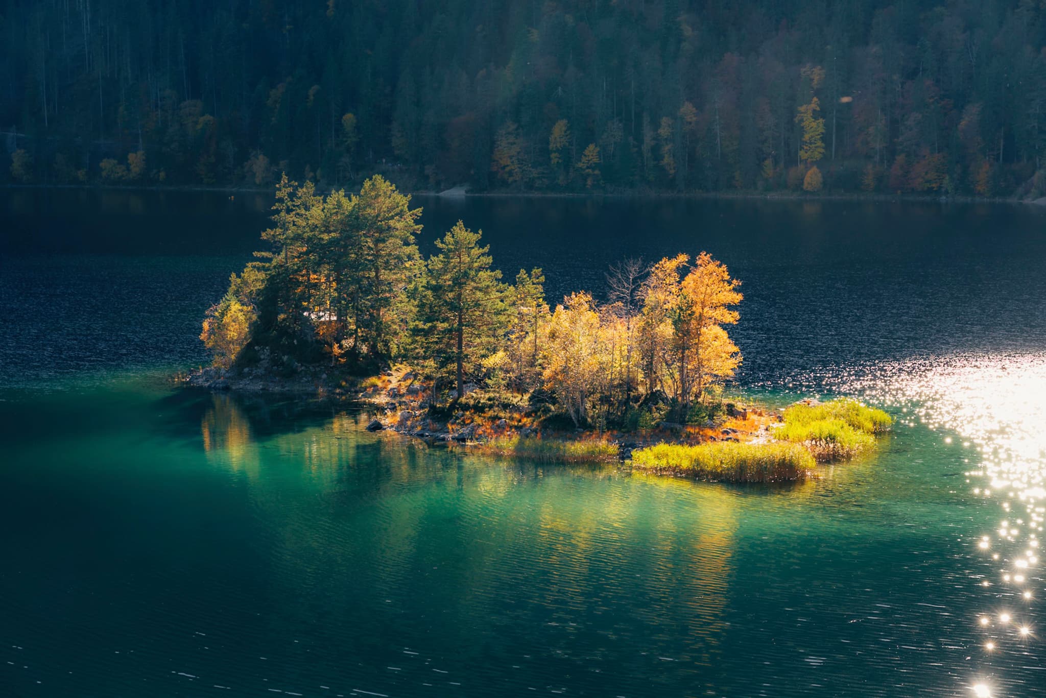 Small tree-covered island glowing with autumn colors in clear turquoise lake with sparkling sunlight on the water