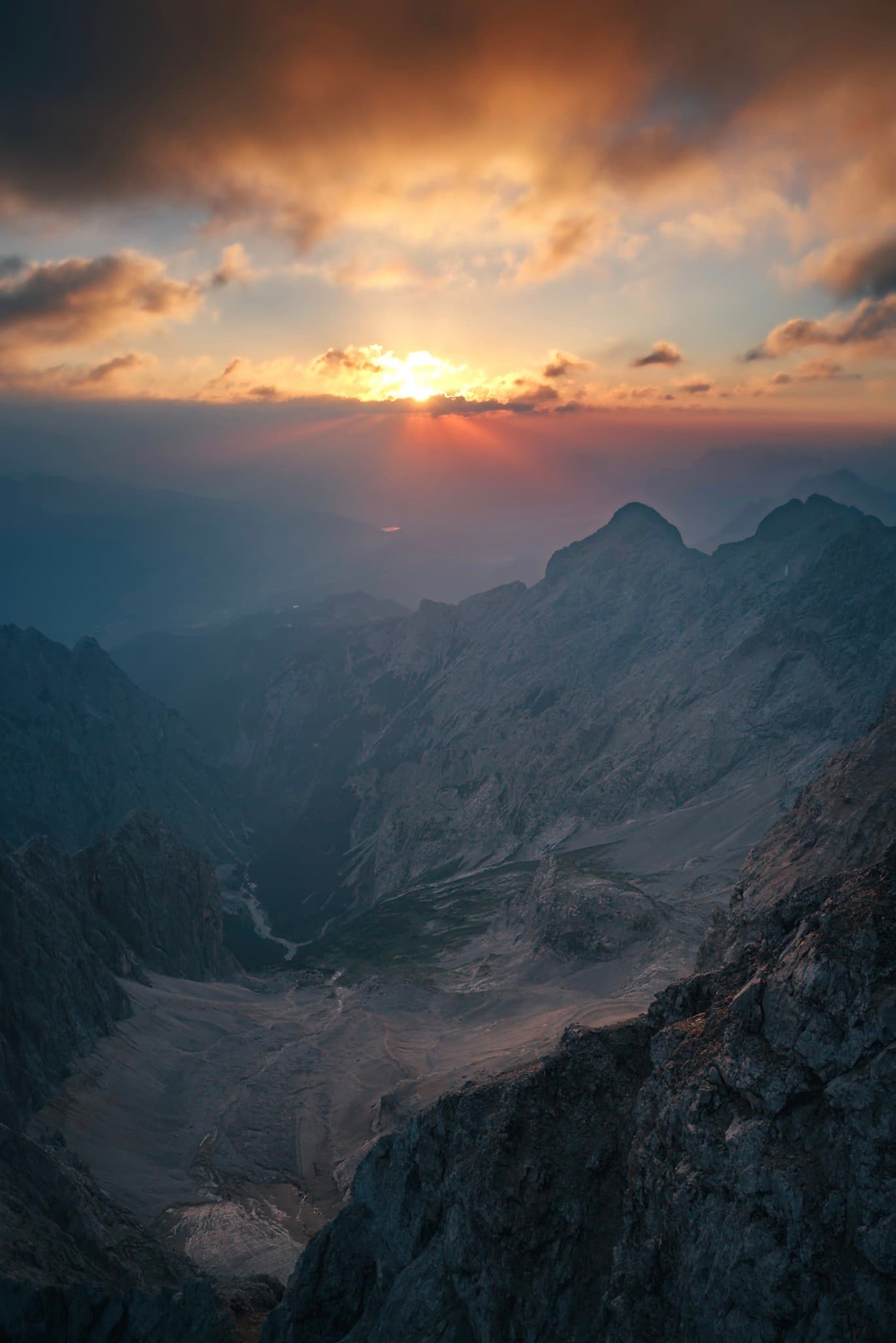 Sun setting behind rugged mountain peaks, casting warm light and shadows into a hazy rocky valley below
