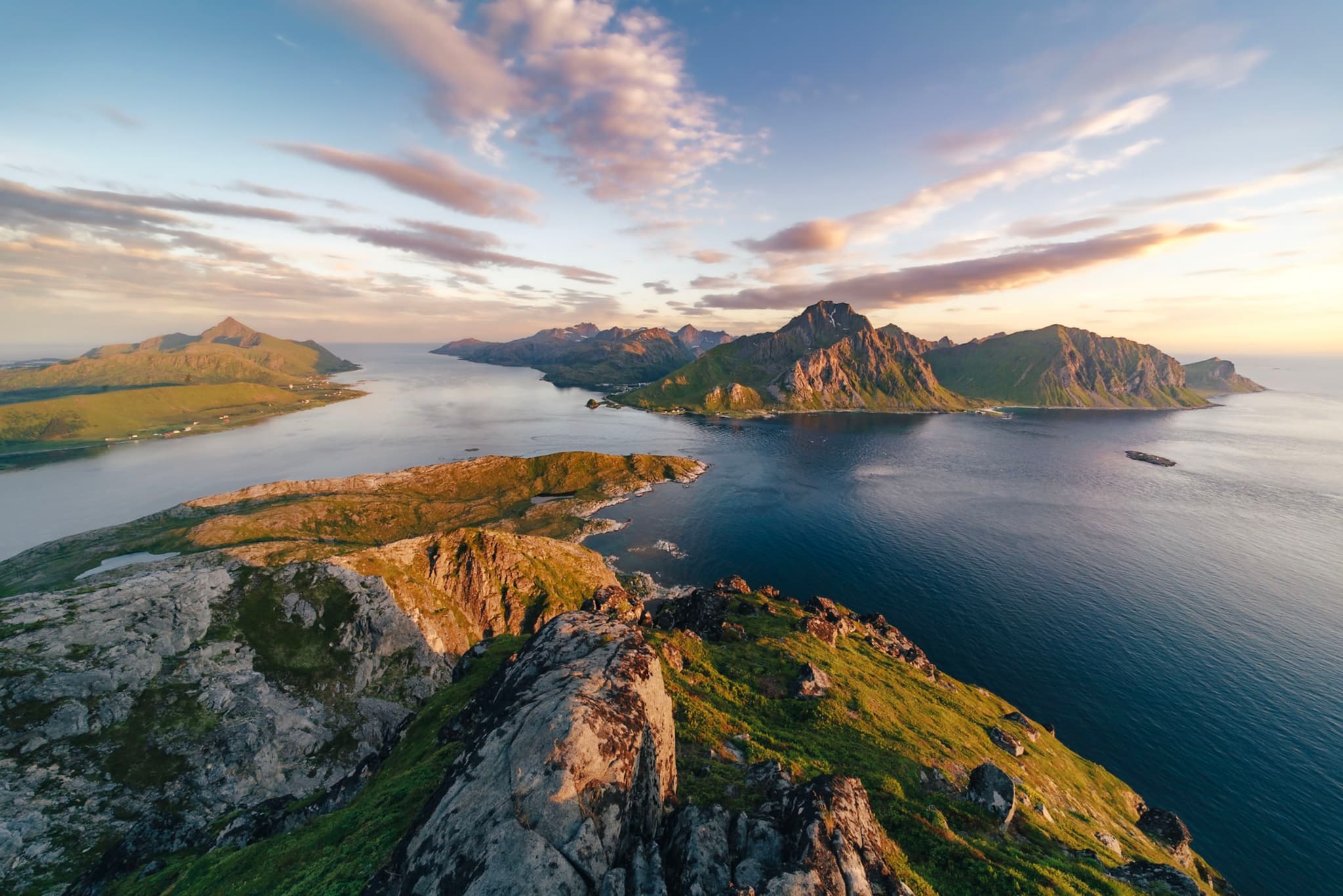 A breathtaking aerial view of a coastal landscape with rugged mountains, calm blue waters, and a partly cloudy sky during sunset