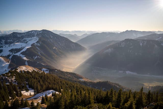 Snow-dusted mountain range with a forested valley filled with soft golden mist under a clear morning sky