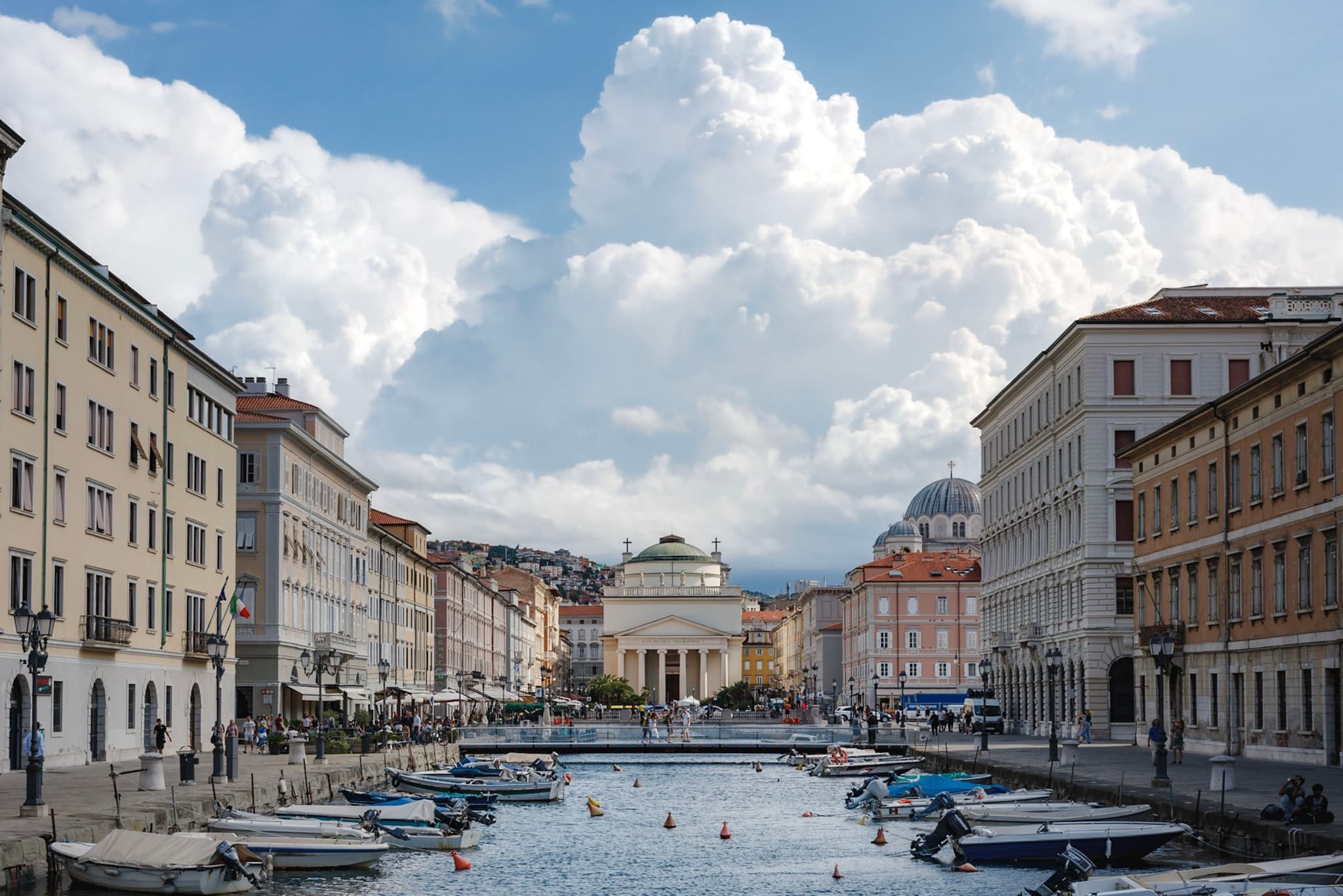 European city canal lined with historic buildings and small boats leading toward a neoclassical domed building under large billowing clouds